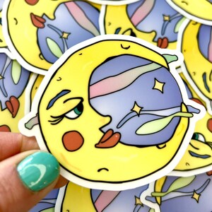 May include: A yellow crescent moon sticker with a face, gazing at a blue planet with a pink and white stripe. The sticker has a white border and is decorated with stars and green accents. The sticker is held by a hand with a teal painted nail.