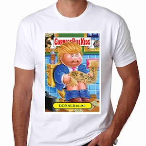 May include: A white t-shirt with a Garbage Pail Kids trading card featuring a cartoon image of Donald Trump as a Cabbage Patch Kid. The card has a yellow background with the text "Garbage Pail Kids" and "Donald Dump". The cartoon image shows Donald Trump sitting on a toilet holding a document that says "We the People".