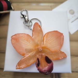 May include: A resin orchid flower keyring in shades of orange and maroon. The flower has detailed veining and a silver-coloured clasp. The keyring is set on a white surface.