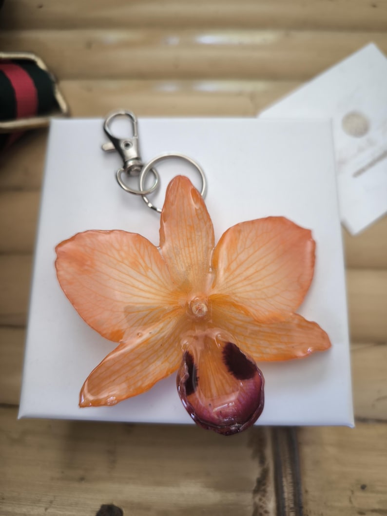 May include: A resin orchid flower keyring in shades of orange and maroon. The flower has detailed veining and a silver-coloured clasp. The keyring is set on a white surface.