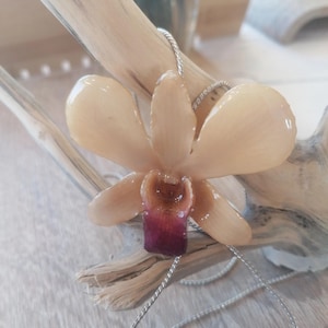 May include: A resin orchid flower pendant necklace with a silver chain. The flower has pale peach petals and a deep burgundy centre. The necklace is displayed on a piece of driftwood.
