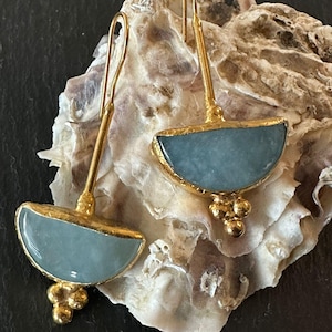 Op de afbeelding: Een paar goudkleurige oorbellen met een lichtblauwe, halfronde steen. De oorbellen hebben een lange, dunne staaf en een kleine cluster van goudkleurige kralen onder de steen. Ze worden gepresenteerd op een getextureerd, gebroken witte ondergrond.