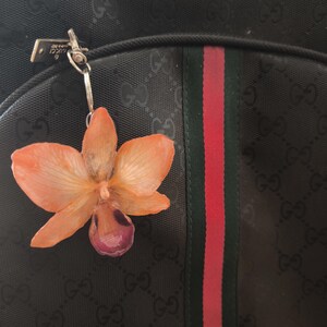 May include: A black handbag with a floral keychain. The keychain is an orange and purple orchid. The handbag has a repeating pattern and a red and green stripe. The bag is a dark colour.