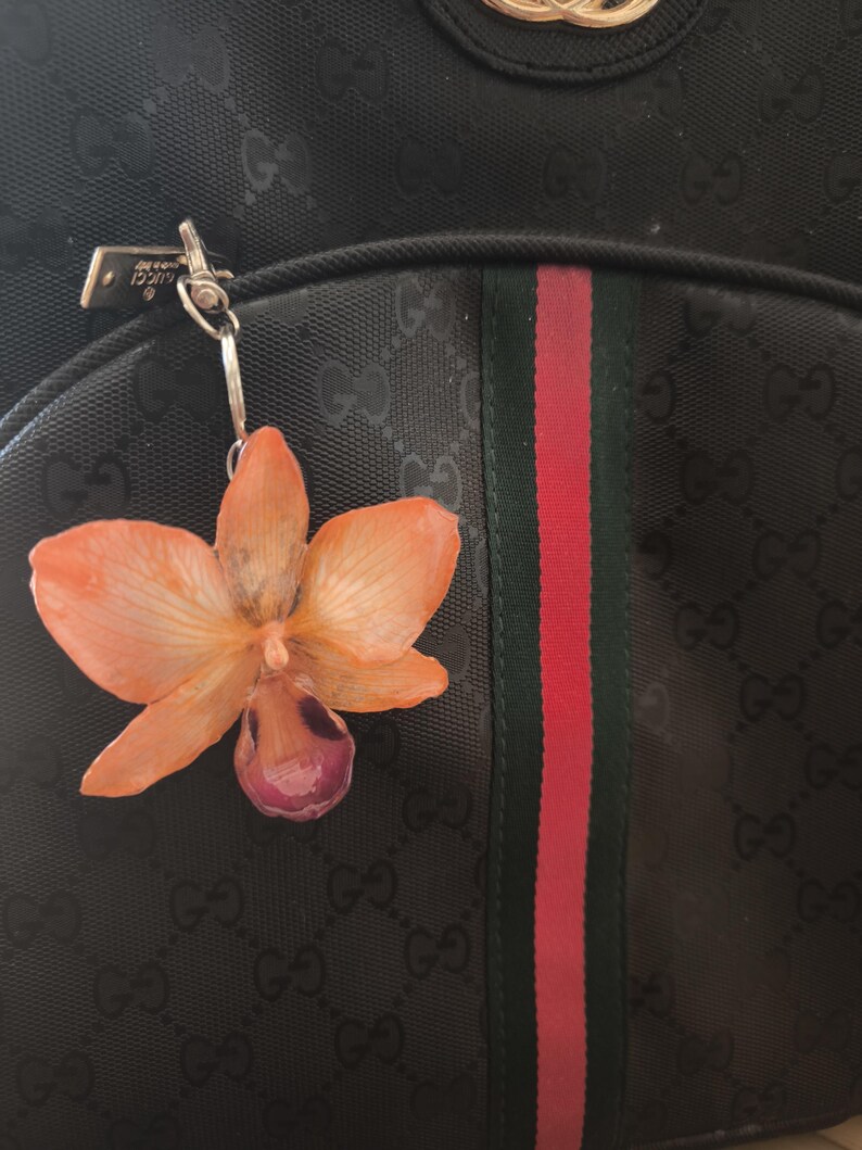 May include: A black handbag with a floral keychain. The keychain is an orange and purple orchid. The handbag has a repeating pattern and a red and green stripe. The bag is a dark colour.