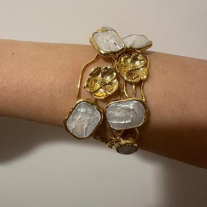 May include: Gold-toned bracelet featuring irregular, pearlescent stones and floral accents. The bracelet's design incorporates organic shapes and textures, creating a unique and elegant accessory. The bracelet is a statement piece.