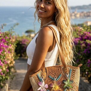 May include: A woven tote bag with leather detailing and floral decorations. The bag has a light brown woven body, a leather top with embossed patterns, and decorative flowers. It has brown leather handles and is being held by a person.