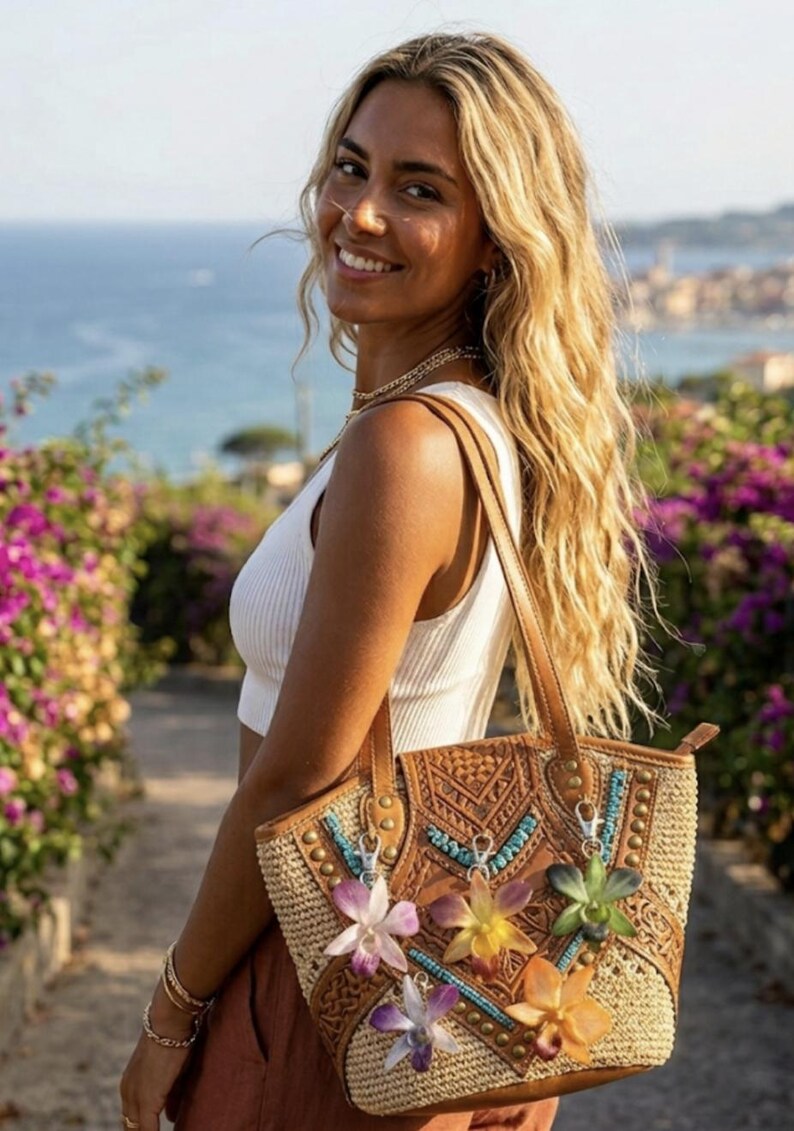 May include: A woven tote bag with leather detailing and floral decorations. The bag has a light brown woven body, a leather top with embossed patterns, and decorative flowers. It has brown leather handles and is being held by a person.