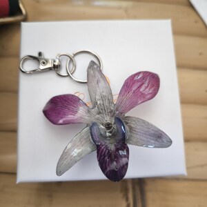 May include: A key ring with a pressed orchid flower, showcasing purple and grey petals. The flower is attached to a silver-coloured metal clasp and ring. The key ring is on a white surface.
