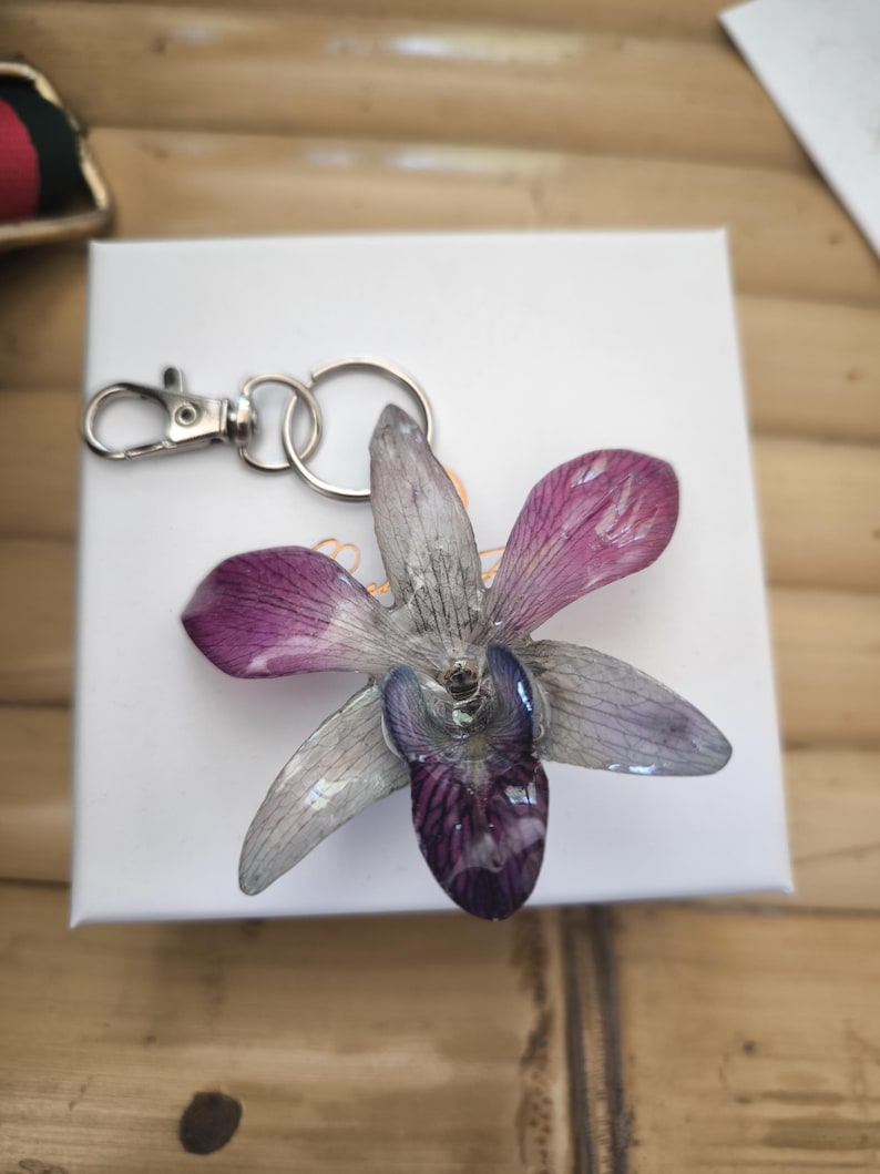May include: A key ring with a pressed orchid flower, showcasing purple and grey petals. The flower is attached to a silver-coloured metal clasp and ring. The key ring is on a white surface.