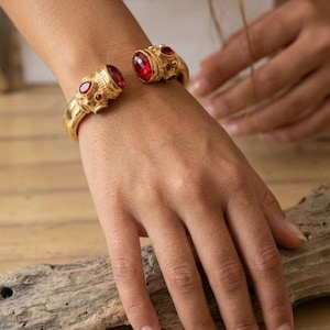 Könnte beinhalten: Ein goldfarbenes Armband in Manschettenform mit zwei großen, ovalen roten Steinen an den offenen Enden, akzentuiert durch kleinere, runde rote Steine, die in das Metallband eingelassen sind. Das Metall hat eine texturierte, matte Oberfläche.
