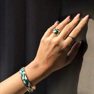 May include: A gold-coloured bracelet and ring set featuring a green and white enamel design. The ring is on a finger, and the bracelet is on a wrist. The jewellery has a bold, geometric pattern.