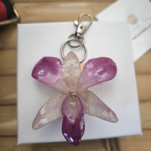 May include: A handmade keychain with a preserved orchid flower in purple and white, set in clear resin. It has a silver-coloured metal clasp and keyring. The keychain is on a white surface, with a wooden background.