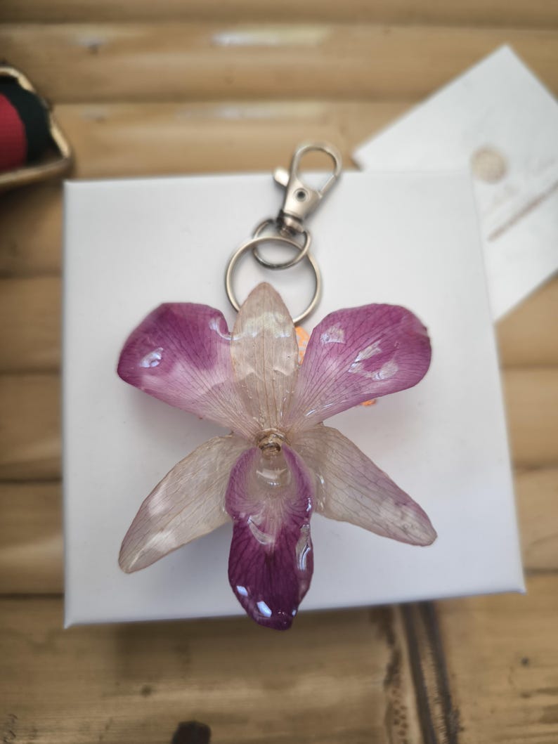 May include: A handmade keychain with a preserved orchid flower in purple and white, set in clear resin. It has a silver-coloured metal clasp and keyring. The keychain is on a white surface, with a wooden background.