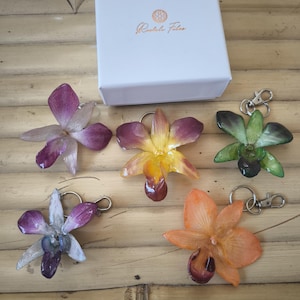 May include: Orchid flower keyrings in shades of purple, orange, yellow, and green, each with a silver clasp. A white box with the text "Rachelle Felas" is visible in the background. The keyrings are arranged on a wooden surface.