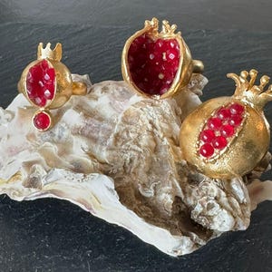 Può includere: Tre anelli dorati con design a forma di melograno. Uno presenta un melograno intero, un altro un melograno tagliato a metà e il terzo un melograno con una corona. I melograni sono riempiti di pietre preziose rosse.