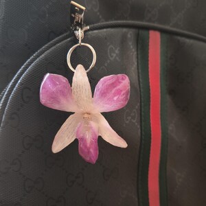 May include: A flower-shaped keyring in shades of pink and white, attached to a black backpack. The backpack features a red and green stripe and a repeating pattern. The keyring is silver.