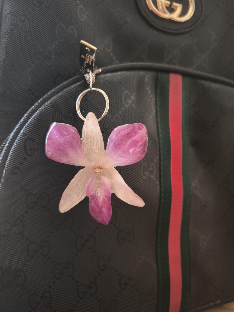 May include: A flower-shaped keyring in shades of pink and white, attached to a black backpack. The backpack features a red and green stripe and a repeating pattern. The keyring is silver.