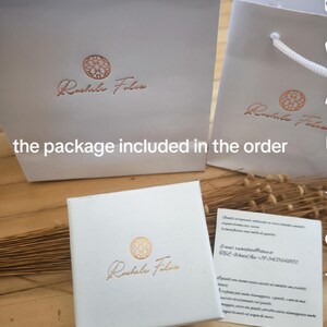 May include: White gift bags and a box with the gold logo "Rachele Foliva". A small card with text is also present. The items are arranged on a wooden surface.