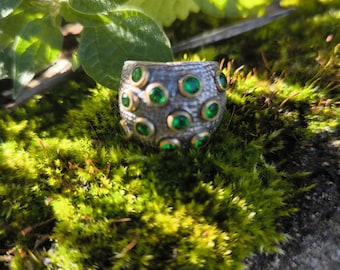 May include: A silver ring with a textured band, adorned with numerous round, green gemstones set in gold-colored bezels. The ring is displayed on a bed of vibrant green moss, with green leaves in the background.