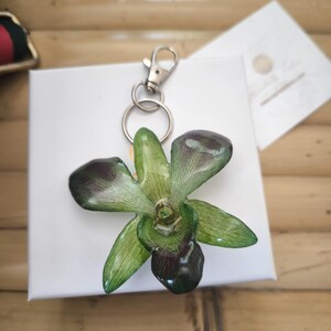 May include: A handmade orchid flower keychain in green and dark purple hues, attached to a silver-tone metal clasp and key ring. The floral accessory is displayed on a white surface. The keychain is a unique gift idea.