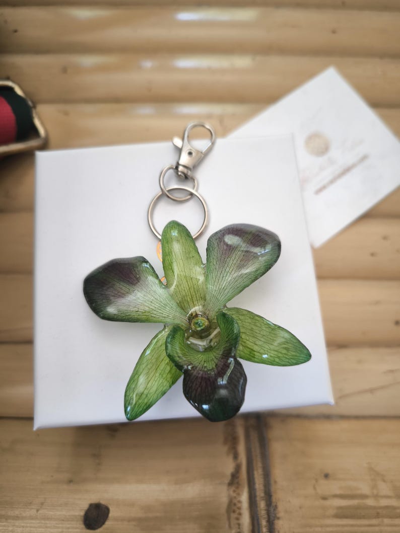 May include: A handmade orchid flower keychain in green and dark purple hues, attached to a silver-tone metal clasp and key ring. The floral accessory is displayed on a white surface. The keychain is a unique gift idea.