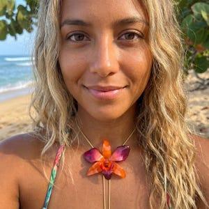 Peut inclure: Une femme porte un collier avec un grand pendentif en forme d'orchidée orange et violet. Le collier a une fine chaîne dorée. La femme est sur une plage avec l'océan en arrière-plan.