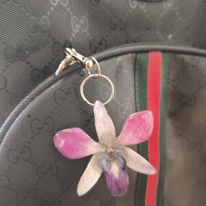 May include: A pressed flower keyring featuring a pink and purple orchid. The keyring is attached to a black backpack with a green and red stripe. The backpack has a repeating pattern.