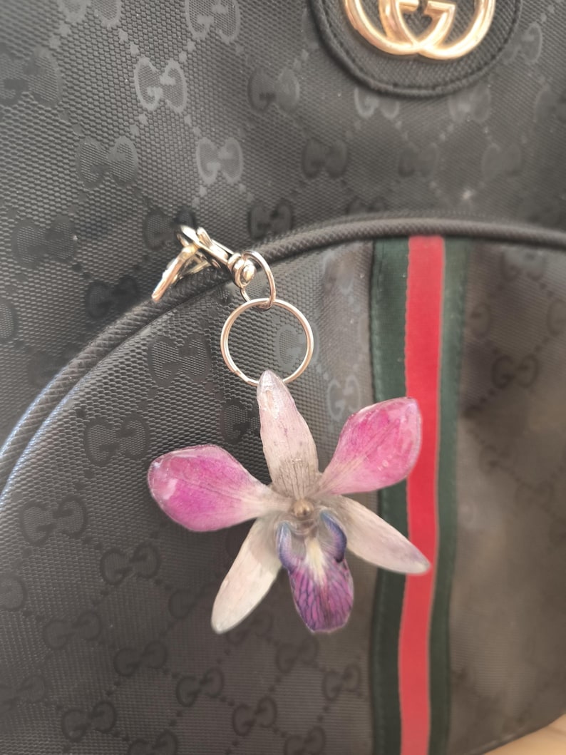May include: A pressed flower keyring featuring a pink and purple orchid. The keyring is attached to a black backpack with a green and red stripe. The backpack has a repeating pattern.