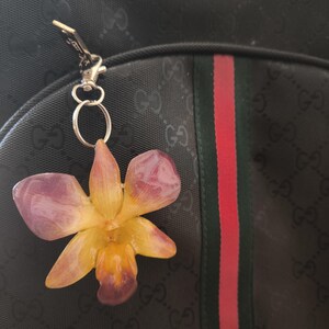 May include: A flower-shaped keyring featuring a purple and yellow orchid design. The keyring is attached to a black backpack with a green and red stripe. The backpack has a repeating pattern of the letters "G" and "O".