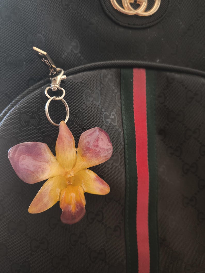May include: A flower-shaped keyring featuring a purple and yellow orchid design. The keyring is attached to a black backpack with a green and red stripe. The backpack has a repeating pattern of the letters "G" and "O".