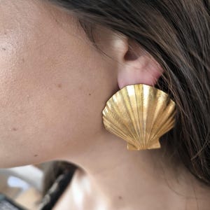 Peut inclure: Une grande boucle d'oreille dorée en forme de coquillage. La boucle d'oreille est portée sur l'oreille et présente une surface texturée avec des nervures détaillées. La boucle d'oreille est une pièce maîtresse, ajoutant une touche d'élégance côtière.