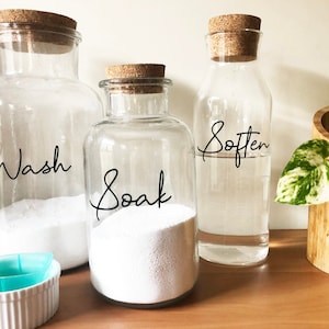 May include: Three glass jars with cork lids labelled "Wash", "Soak", and "Soften". The jars are filled with white powder and clear liquid. The jars are sitting on a wooden surface.