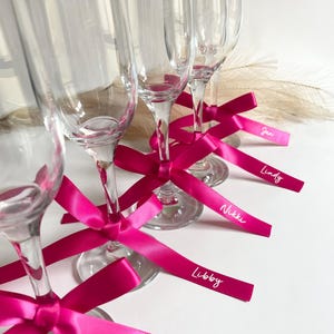 Pu&ograve; includere: Fl&ucirc;te di champagne trasparenti con nastri rosa acceso legati attorno agli steli. Ogni nastro ha un nome stampato in bianco. Le fl&ucirc;te sono disposte su una superficie bianca, con alcune piume decorative sullo sfondo.