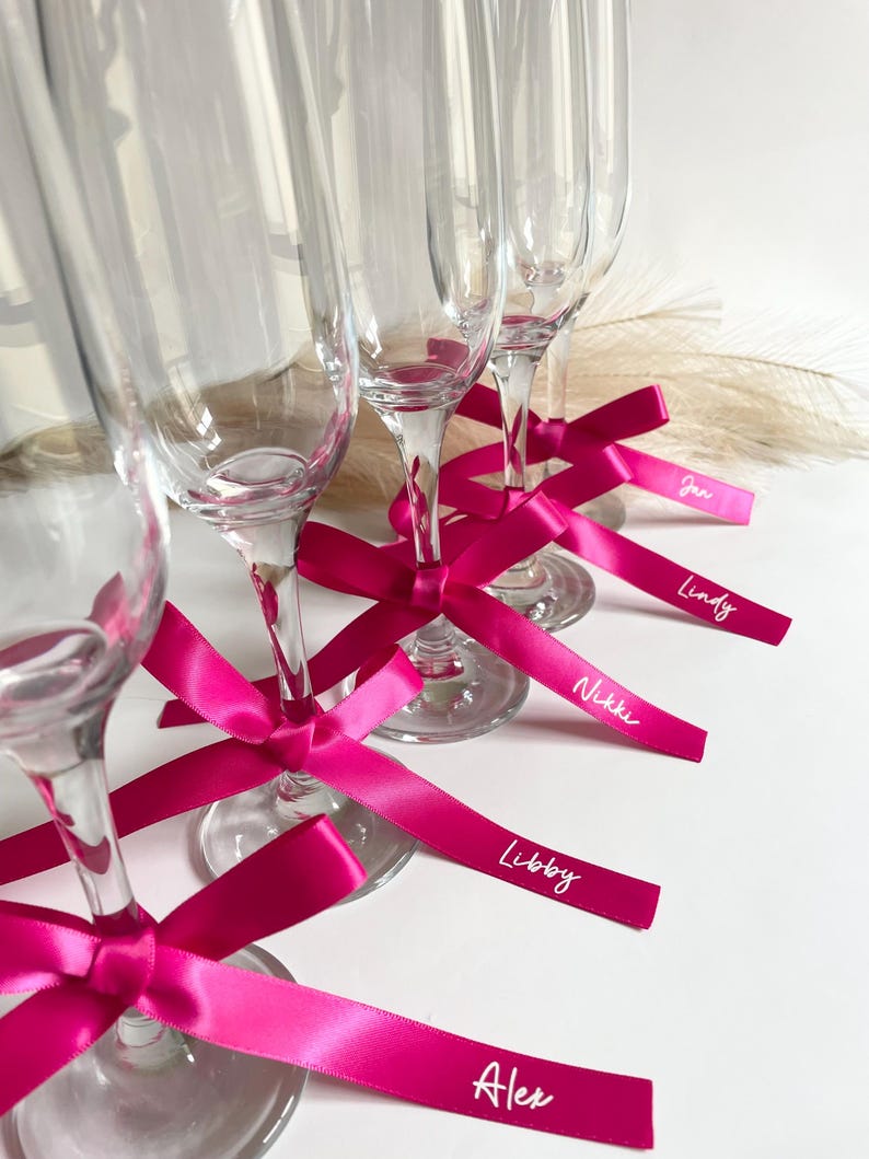 Pu&ograve; includere: Fl&ucirc;te di champagne trasparenti con nastri rosa acceso legati attorno agli steli. Ogni nastro ha un nome stampato in bianco. Le fl&ucirc;te sono disposte su una superficie bianca, con alcune piume decorative sullo sfondo.