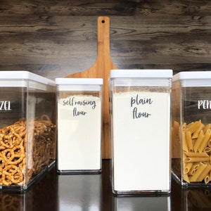 May include: Four clear plastic containers with white lids, labelled "Pretzel", "Self-raising Flour", "Plain Flour", and "Penne". The containers are filled with pretzels, flour, and pasta.