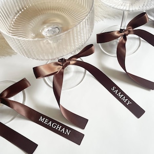 Pu&ograve; includere: Bicchieri da champagne in vetro trasparente con etichette con nomi in nastro di raso marrone. Il nastro &egrave; legato attorno allo stelo del bicchiere e presenta nomi in lettere bianche. Su una superficie bianca.