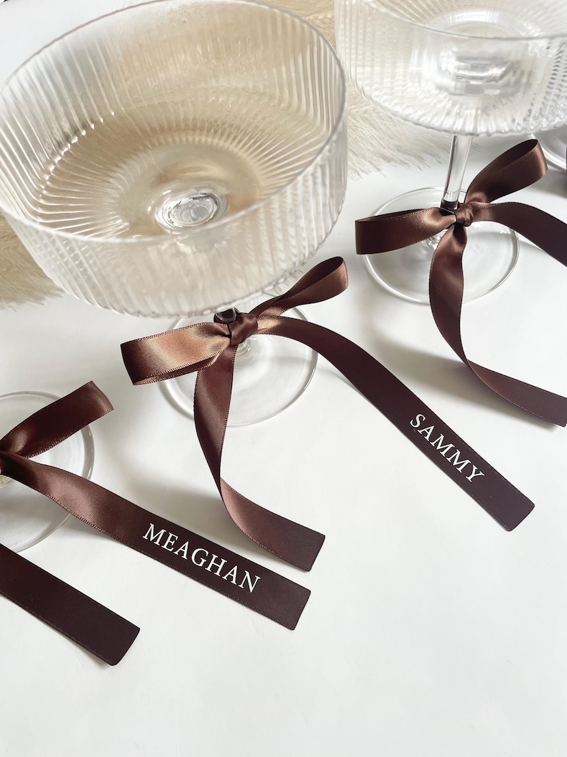 Pu&ograve; includere: Bicchieri da champagne in vetro trasparente con etichette con nomi in nastro di raso marrone. Il nastro &egrave; legato attorno allo stelo del bicchiere e presenta nomi in lettere bianche. Su una superficie bianca.