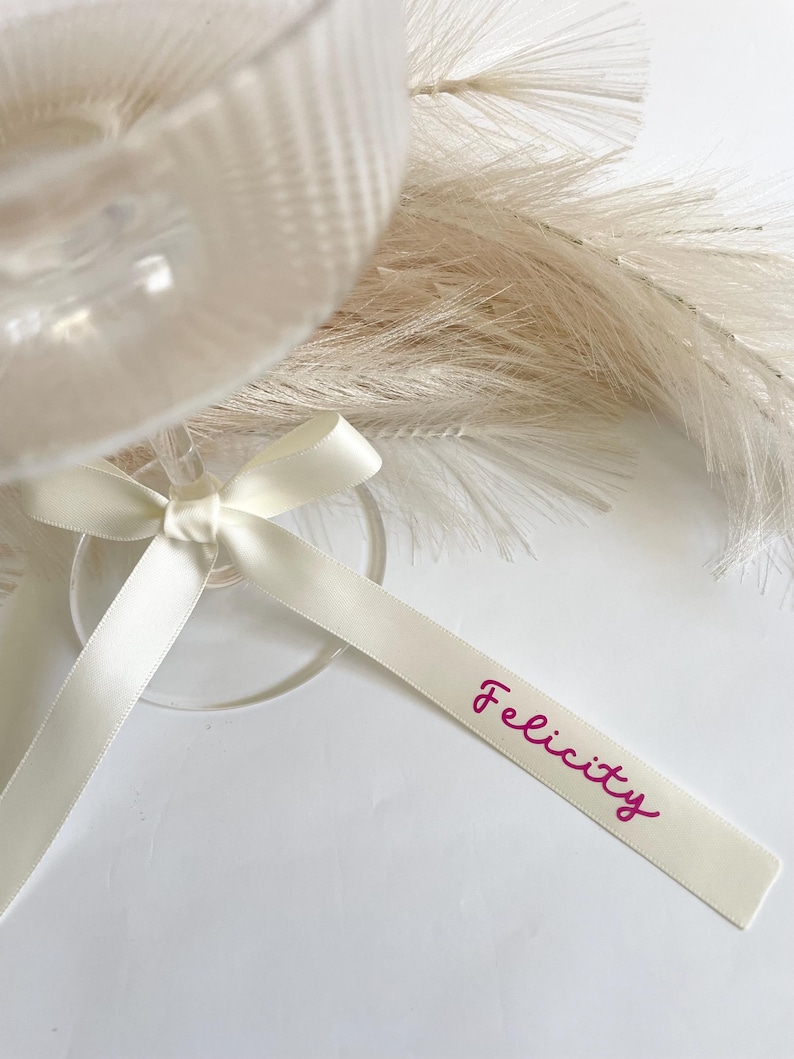 Pu&ograve; includere: Un bicchiere da vino trasparente con un nastro di raso bianco legato attorno allo stelo. Il nastro ha il nome "Felicity" stampato in magenta. Lo sfondo include elementi decorativi bianchi e soffici.