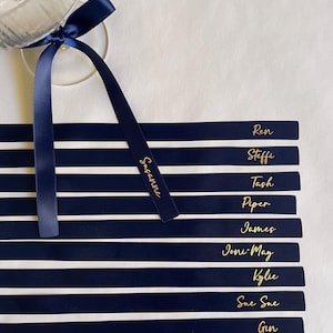 Pu&ograve; includere: Segnaposto a righe blu navy e bianche con scritte dorate. Ogni biglietto riporta un nome, come "Ren" e "Steffi". Un nastro blu navy &egrave; legato a un calice. Il nastro riporta il nome "Susanne" in lettere dorate.
