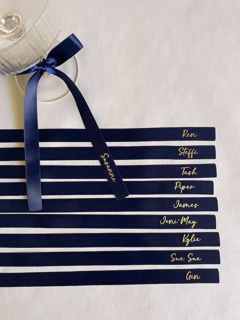 Pu&ograve; includere: Segnaposto a righe blu navy e bianche con scritte dorate. Ogni biglietto riporta un nome, come "Ren" e "Steffi". Un nastro blu navy &egrave; legato a un calice. Il nastro riporta il nome "Susanne" in lettere dorate.