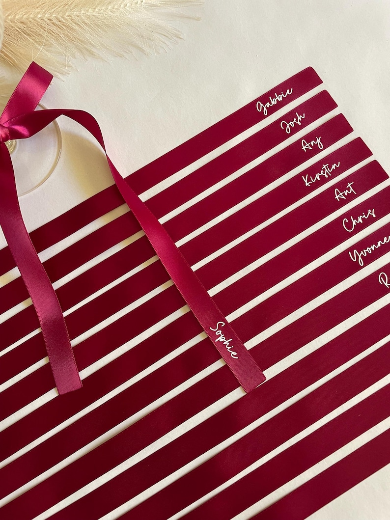 Pu&ograve; includere: Segnaposto bordeaux con nomi bianchi, come Sophie e Josh. Un nastro bordeaux abbinato &egrave; legato a fiocco. Le carte sono disposte in diagonale, perfette per un matrimonio o un evento.