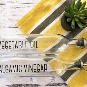 May include: Two clear glass bottles with black labels and pour spouts. The top bottle is labelled "VEGETABLE OIL" and the bottom bottle is labelled "BALSAMIC VINEGAR".