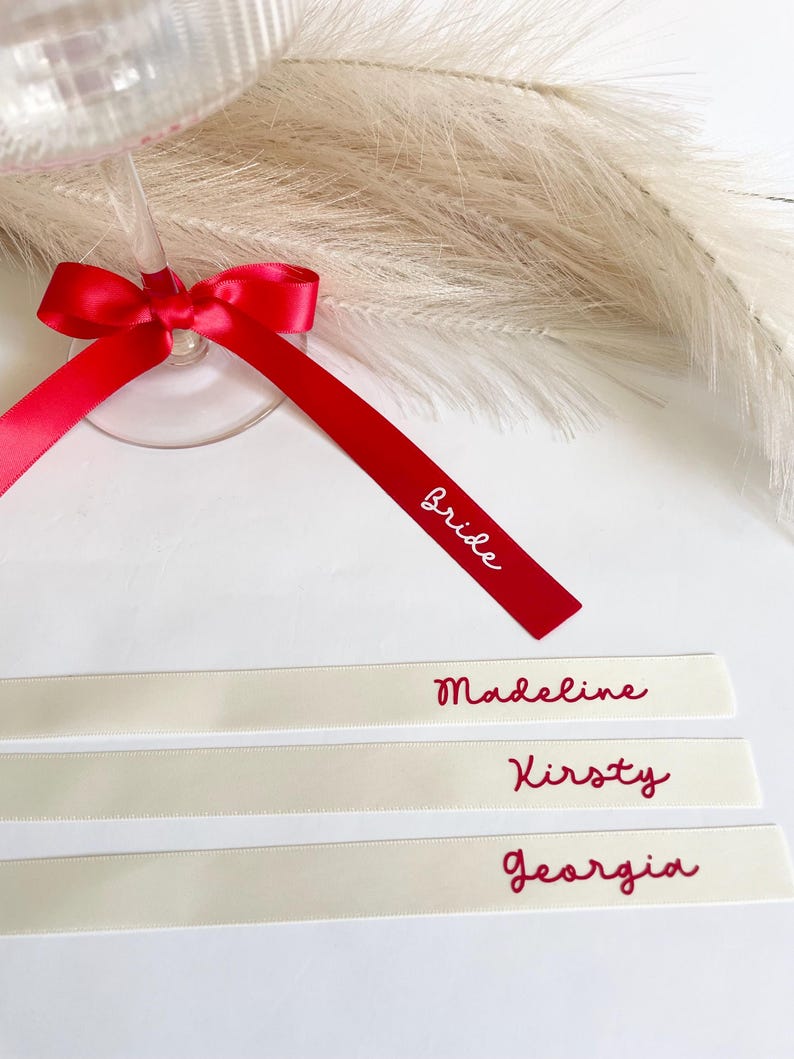 Pu&ograve; includere: Un calice da champagne con un nastro rosso legato a fiocco e la parola "Bride". Sotto, tre nastri color avorio con i nomi "Madeline", "Kirsty" e "Georgia" in rosso. Perfetto per matrimoni.