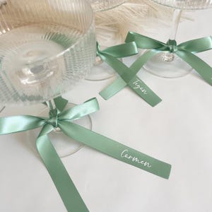 Pu&ograve; includere: Eleganti calici da champagne con nastri di raso verde salvia legati attorno agli steli. I nastri hanno nomi stampati in corsivo bianco. I calici sono su una superficie bianca, perfetti per un matrimonio.