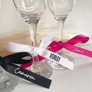 Pu&ograve; includere: Primo piano di calici da champagne con etichette nominative personalizzate con nastro. I nastri sono neri, bianchi e fucsia, con nomi come "Cameron" e "Ashley". I calici di vetro trasparente sono ideali per un matrimonio o un evento speciale.