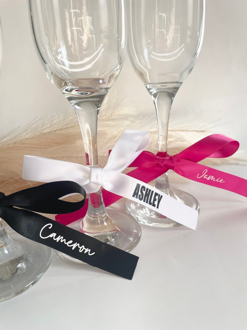 Pu&ograve; includere: Primo piano di calici da champagne con etichette nominative personalizzate con nastro. I nastri sono neri, bianchi e fucsia, con nomi come "Cameron" e "Ashley". I calici di vetro trasparente sono ideali per un matrimonio o un evento speciale.