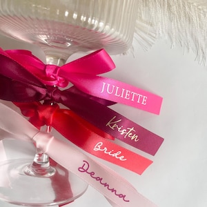 Pu&ograve; includere: Primo piano di un calice di vetro con nastri colorati. I nastri, in varie tonalit&agrave; di rosa e rosso, sono stampati con nomi in oro e bianco. I nomi includono "Juliette", "Kristen", "Bride" e "Deanna".