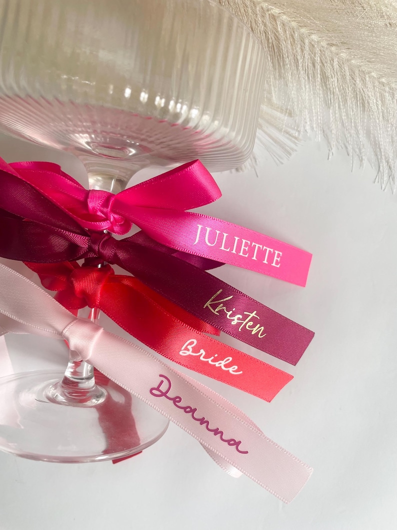 Pu&ograve; includere: Primo piano di un calice di vetro con nastri colorati. I nastri, in varie tonalit&agrave; di rosa e rosso, sono stampati con nomi in oro e bianco. I nomi includono "Juliette", "Kristen", "Bride" e "Deanna".