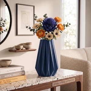Può includere: Un vaso geometrico blu navy contiene un vivace bouquet di fiori arancioni, bianchi e blu. Il vaso è appoggiato su un piano di lavoro maculato con libri e un tavolo di legno. Uno specchio rotondo e opere d'arte incorniciate sono sullo sfondo.