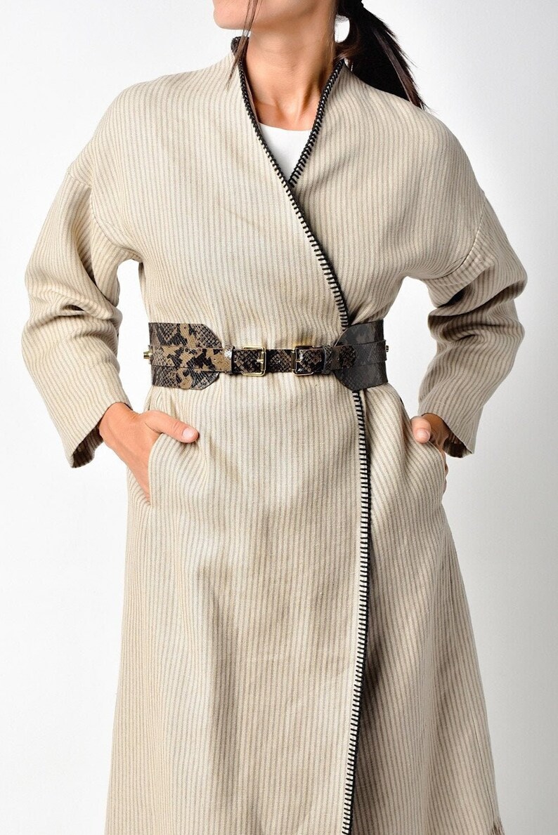 May include: A long, beige striped coat with a black zipper down the front. The coat has a wide, black and brown snakeskin belt with a gold buckle.